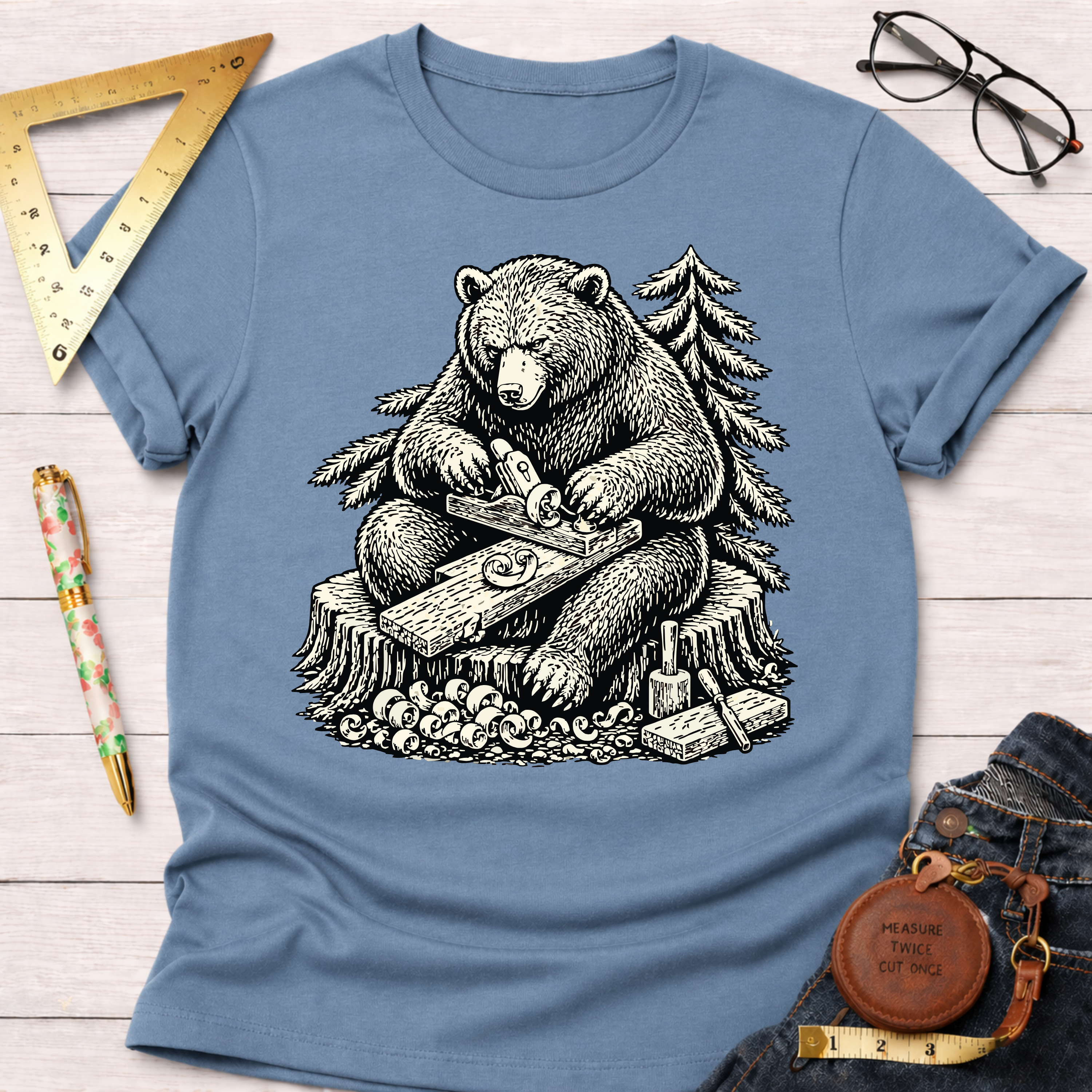 Women's funny woodworking graphic tee shirt for female woodworkers, makers, and DIY builders — Sawdust & Ponytails. Light blue cotton tee with a black and white graphic of a bear hand planing wood on a tree stump.