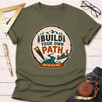 Olive green "Build Your Own Path" t-shirt with a wood slice graphic, mountains, tools, and a river.
