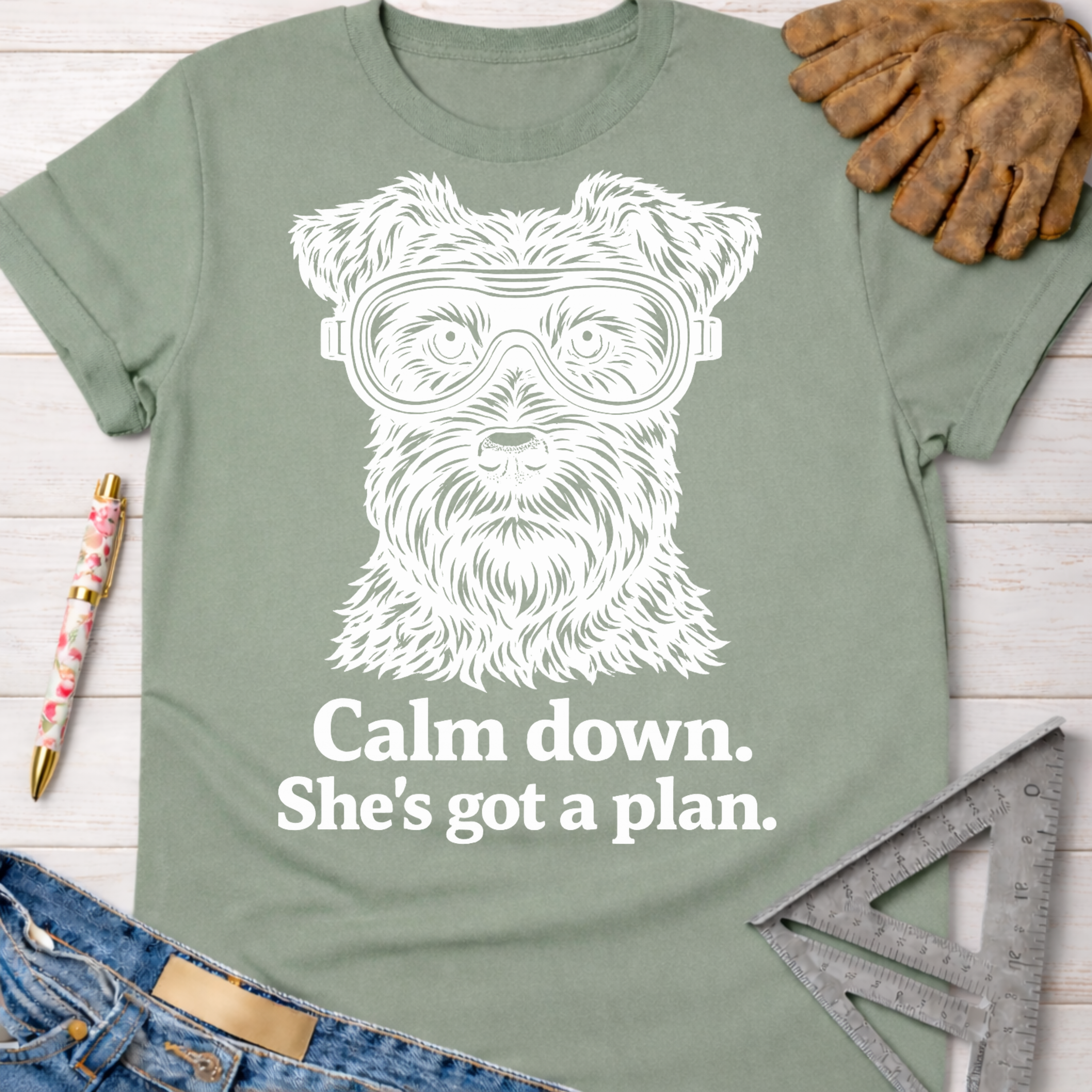 Women's funny woodworking graphic tee shirt for female woodworkers, makers, and DIY builders — Sawdust & Ponytails. Sage green cotton t-shirt with a white graphic of a dog wearing safety goggles and "Calm down. She's got a plan." text.