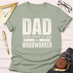 Sage green "Dad The Man The Myth The Legend Woodworker" t-shirt on a white wood surface.