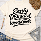 Women's funny woodworking graphic tee shirt for female woodworkers, makers, and DIY builders — Sawdust & Ponytails. Cream t-shirt with "Easily Distracted by Wood & Tools" in black script.