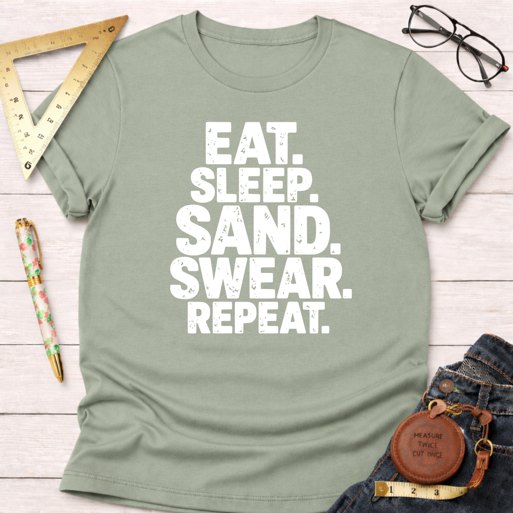 Sage green "Eat Sleep Sand Swear Repeat" t-shirt with rolled sleeves, beside jeans, ruler, pen, and glasses.