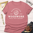 Dusty rose t-shirt with "HANDCRAFTED WOODWORK BUILT NOT BOUGHT" graphic, styled with jeans and tools.