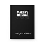 Maker's Journal hardcover with matte black wraparound cover, closed on a workbench