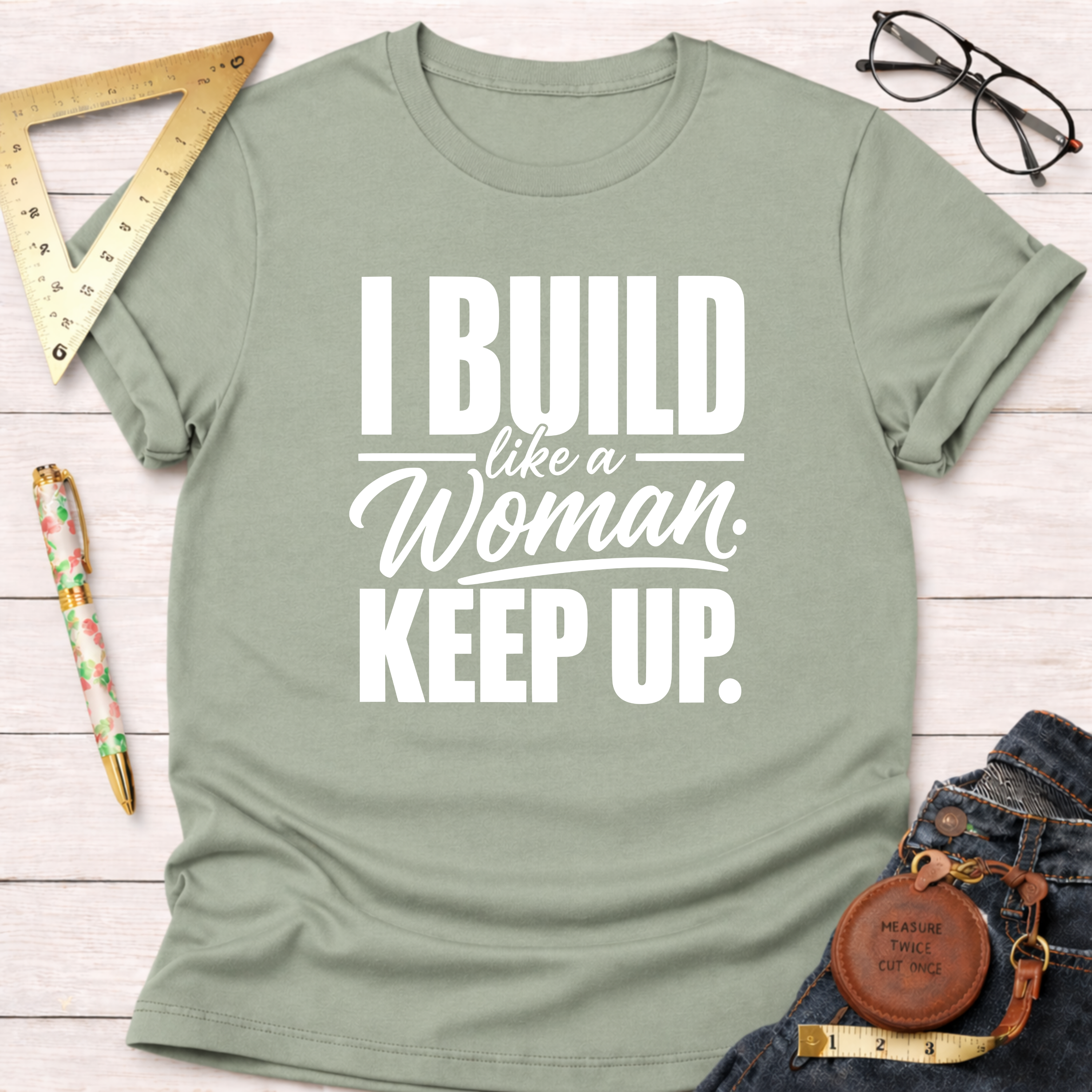 Sage green cotton t-shirt with "I Build Like A Woman. Keep Up." in white text, styled on a white wood surface.