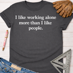 Women's funny woodworking graphic tee shirt for female woodworkers, makers, and DIY builders — Sawdust & Ponytails. Dark gray cotton tee with "I like working alone more than I like people" in white text, styled with jeans, work glove, and ruler.