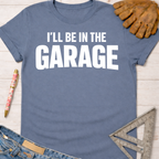 Women's funny woodworking graphic tee shirt for female woodworkers, makers, and DIY builders — Sawdust & Ponytails. Blue cotton t-shirt with "I'LL BE IN THE GARAGE" in white text.