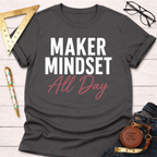 Dark gray "Maker Mindset All Day" t-shirt with white and pink text, folded on a white wood surface.