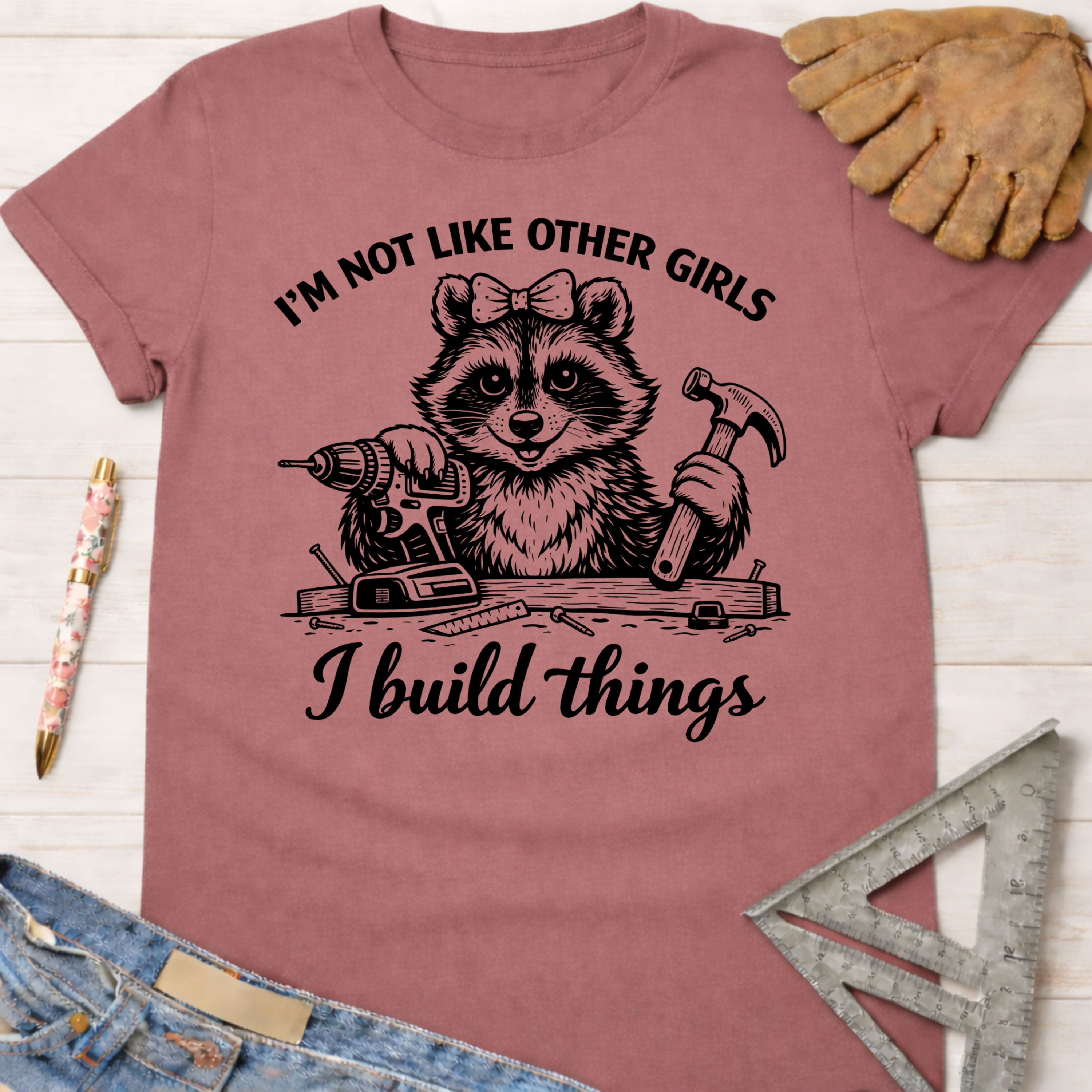 Women's funny woodworking graphic tee shirt for female woodworkers, makers, and DIY builders — Sawdust & Ponytails. Mauve t-shirt with a raccoon holding a drill and hammer, text "I'm not like other girls I build things.