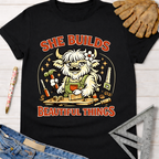 Black cotton tee with "She Builds Beautiful Things" and a cartoon highland cow building. Women's funny woodworking graphic tee shirt for female woodworkers, makers, and DIY builders — Sawdust & Ponytails