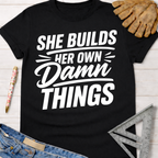 Black cotton tee with "SHE BUILDS HER OWN Damn THINGS" in white text. Women's funny woodworking graphic tee shirt for female woodworkers, makers, and DIY builders — Sawdust & Ponytails.