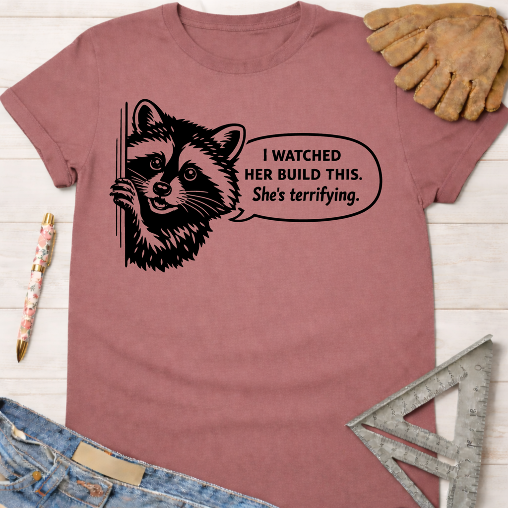 Women's funny woodworking graphic tee shirt for female woodworkers, makers, and DIY builders — Sawdust & Ponytails. Mauve t-shirt with a black graphic of a raccoon and speech bubble "I watched her build this. She's terrifying.