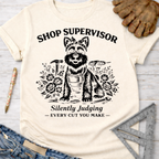 Women's funny woodworking graphic tee shirt for female woodworkers, makers, and DIY builders — Sawdust & Ponytails. Cream t-shirt with a black graphic of a dog in a bandana and apron, surrounded by tools and flowers, with "Shop Supervisor Silently Judging – EVERY CUT YOU MAKE –" text.