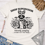 Women's funny woodworking graphic tee shirt for female woodworkers, makers, and DIY builders — Sawdust & Ponytails. White cotton tee with a black graphic of a dog in a bandana and apron, surrounded by tools and flowers, with text "Shop Supervisor Silently Judging Every Cut You Make.