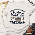 Cream t-shirt with "Never Underestimate An Old Man Who Still Does Woodworking In His Eighties" text and tools.