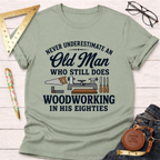 Sage green t-shirt with "Never Underestimate An Old Man Who Still Does Woodworking In His Eighties" text and tools graphic.