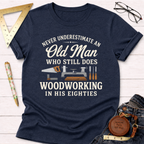 Navy blue heather t-shirt with "Never Underestimate An Old Man Who Still Does Woodworking In His Eighties" text and woodworking tools graphic.
