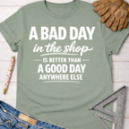 Women's funny woodworking graphic tee shirt for female woodworkers, makers, and DIY builders — Sawdust & Ponytails. Sage green cotton tee with "A BAD DAY in the shop IS BETTER THAN A GOOD DAY ANYWHERE ELSE" text.