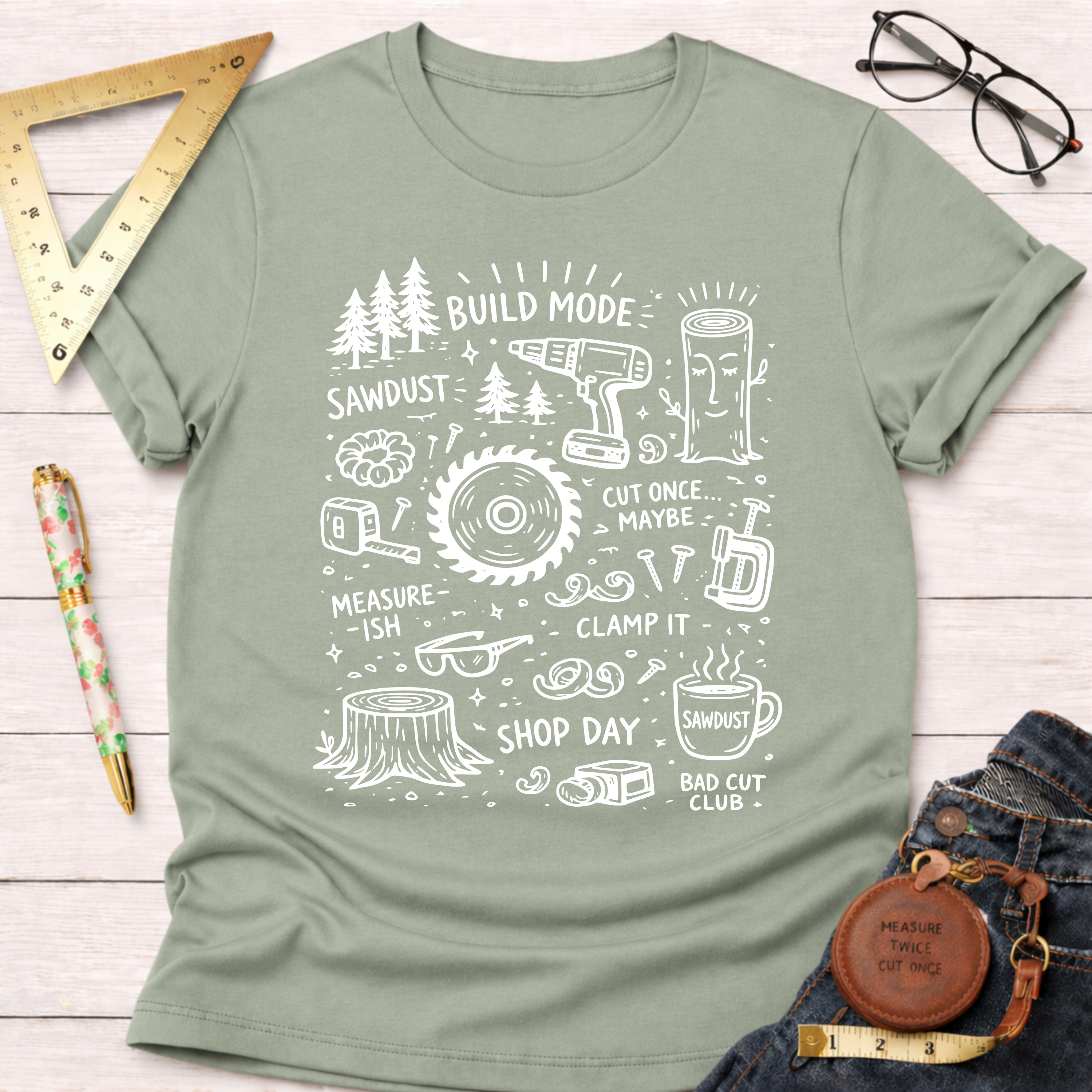 Women's funny woodworking graphic tee shirt for female woodworkers, makers, and DIY builders — Sawdust & Ponytails. Sage green crewneck t-shirt with white "Build Mode" tools and wood-themed doodles.