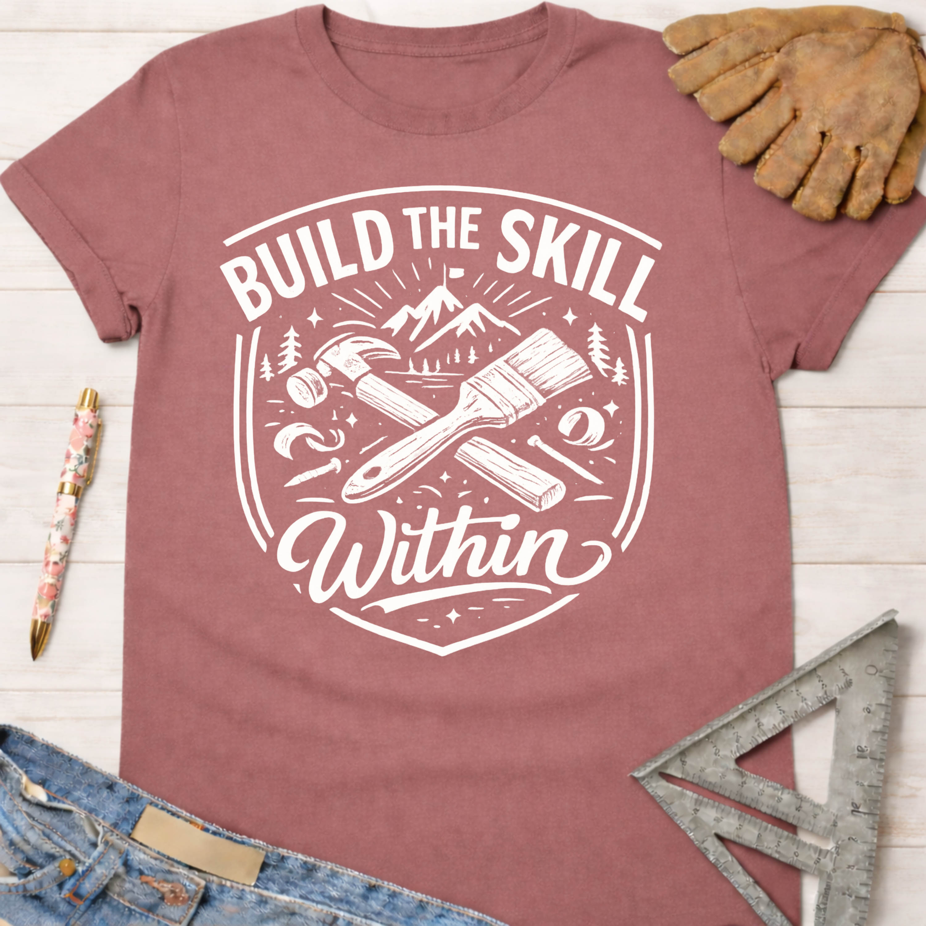 Women's funny woodworking graphic tee shirt for female woodworkers, makers, and DIY builders — Sawdust & Ponytails. Mauve t-shirt with "Build the Skill Within" graphic, hammer, paintbrush, and mountains.