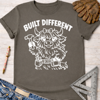 Women's funny woodworking graphic tee shirt for female woodworkers, makers, and DIY builders — Sawdust & Ponytails. Dark gray cotton tee with "Built Different" and a cute highland cow holding a hammer.