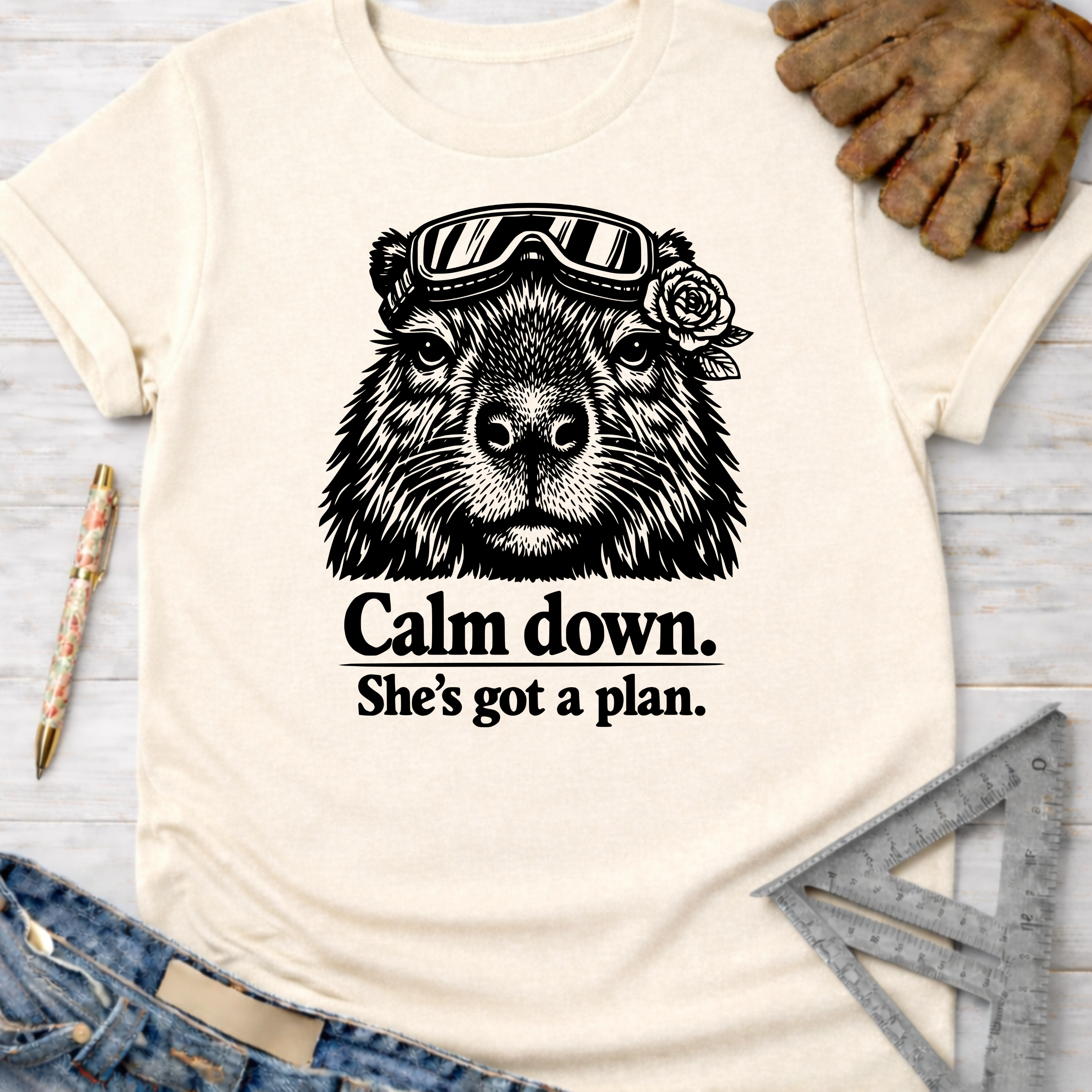Women's funny woodworking graphic tee shirt for female woodworkers, makers, and DIY builders — Sawdust & Ponytails. Cream crewneck t-shirt with a black graphic of a capybara wearing goggles and a rose, with the text "Calm down. She's got a plan.