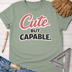 Women's funny woodworking graphic tee shirt for female woodworkers, makers, and DIY builders — Sawdust & Ponytails. Sage green cotton t-shirt with "Cute BUT CAPABLE." graphic, shown with jeans, work glove, and ruler.