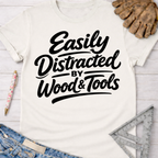 Women's funny woodworking graphic tee shirt for female woodworkers, makers, and DIY builders — Sawdust & Ponytails. Cream t-shirt with "Easily Distracted by Wood & Tools" in black script.