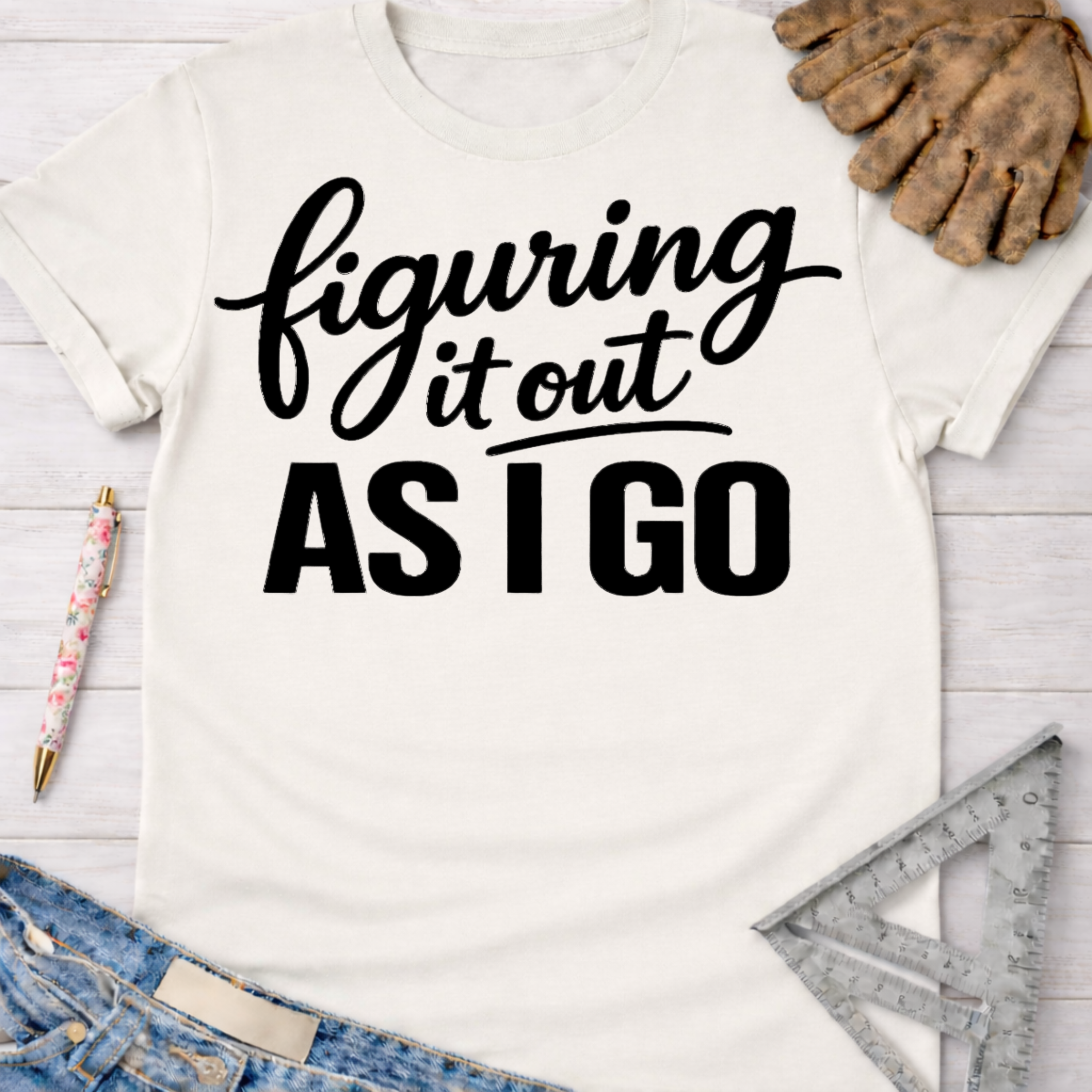 Women's funny woodworking graphic tee shirt for female woodworkers, makers, and DIY builders — Sawdust & Ponytails. Cream t-shirt with "figuring it out AS I GO" in black script and block text, styled with jeans, work gloves, and a ruler.