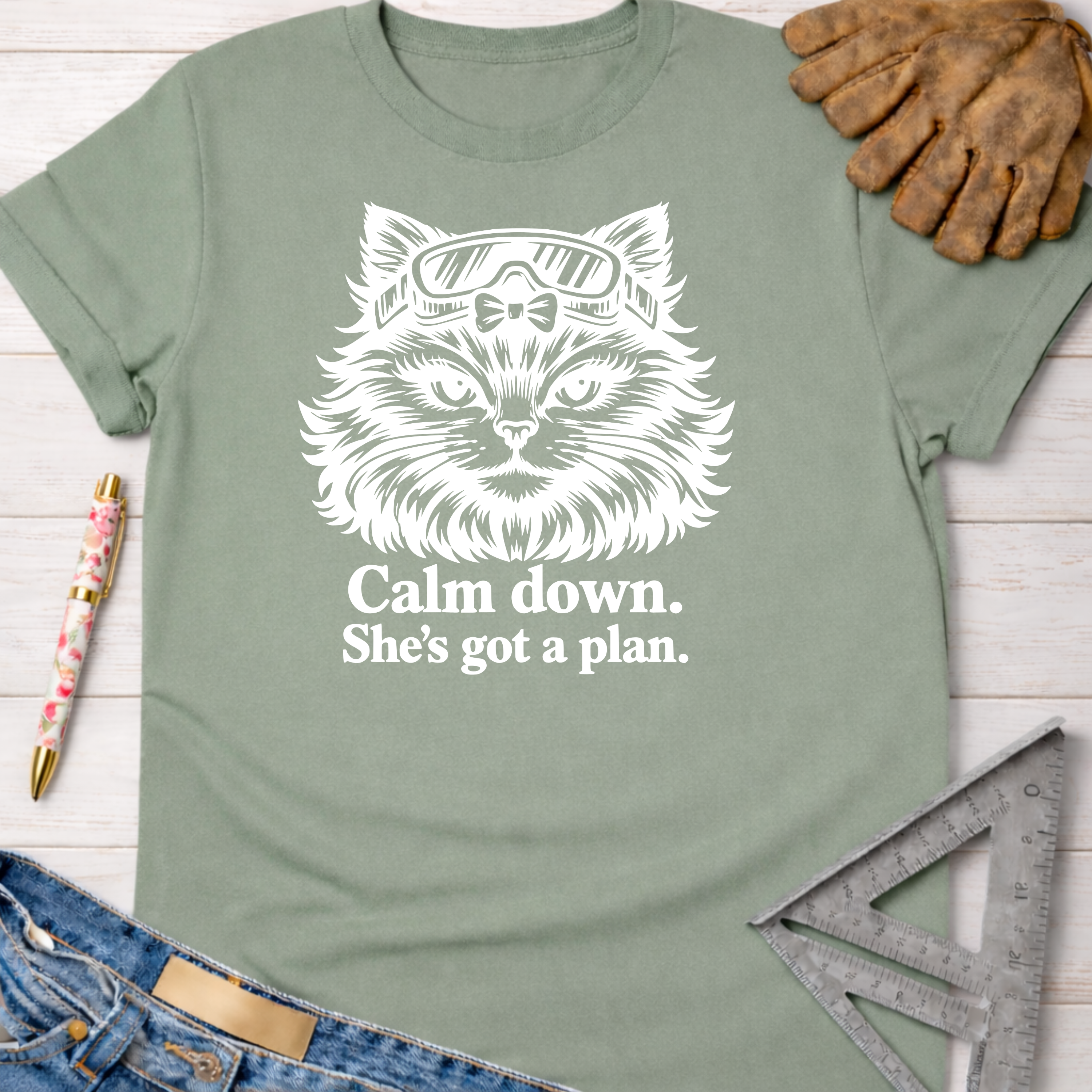 Sage green women's cotton tee with a white graphic of a cat in goggles and text "Calm down. She's got a plan." Women's funny woodworking graphic tee shirt for female woodworkers, makers, and DIY builders — Sawdust & Ponytails