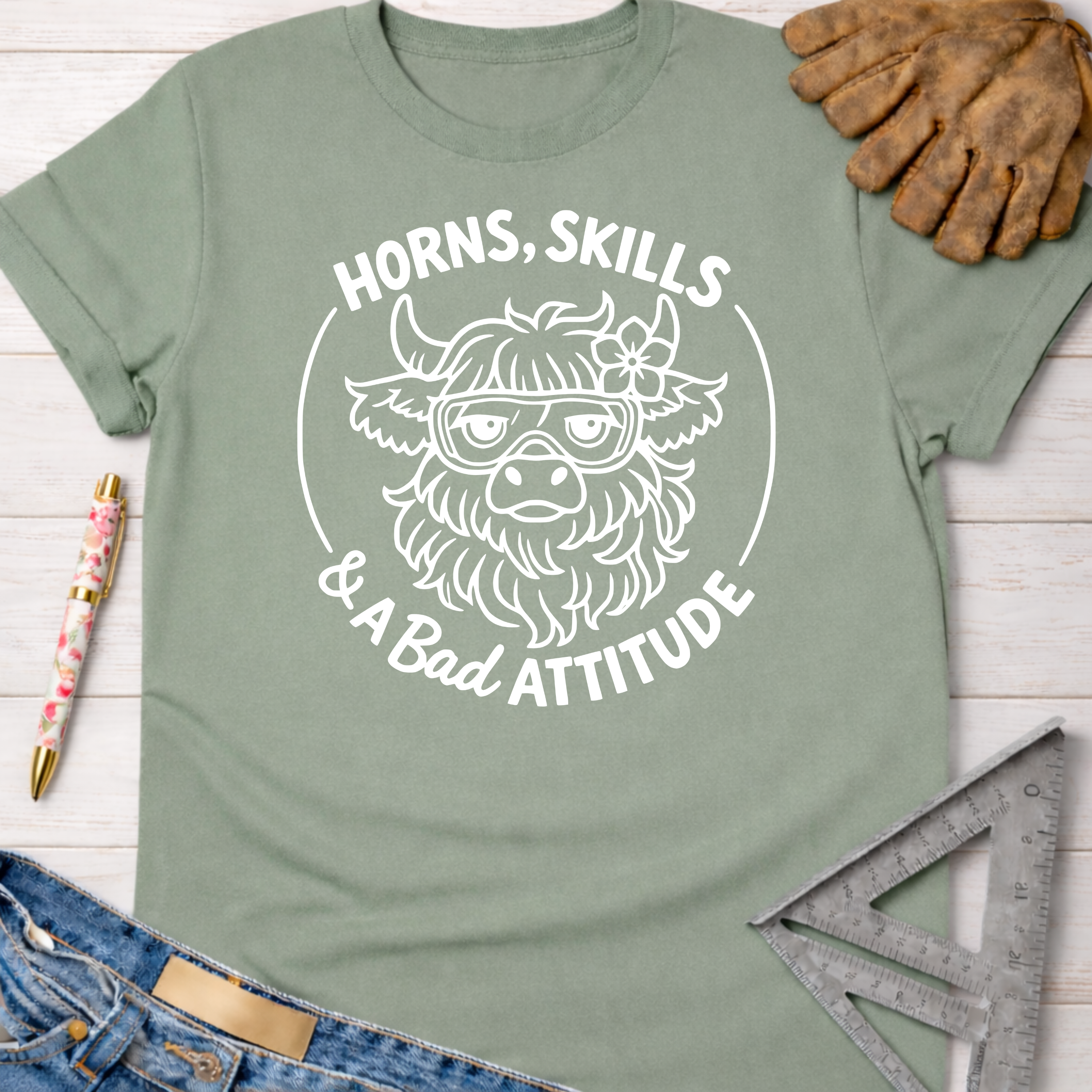 Women's funny woodworking graphic tee shirt for female woodworkers, makers, and DIY builders — Sawdust & Ponytails. Sage green cotton tee with "Horns, Skills & A Bad Attitude" text and a cow wearing goggles.