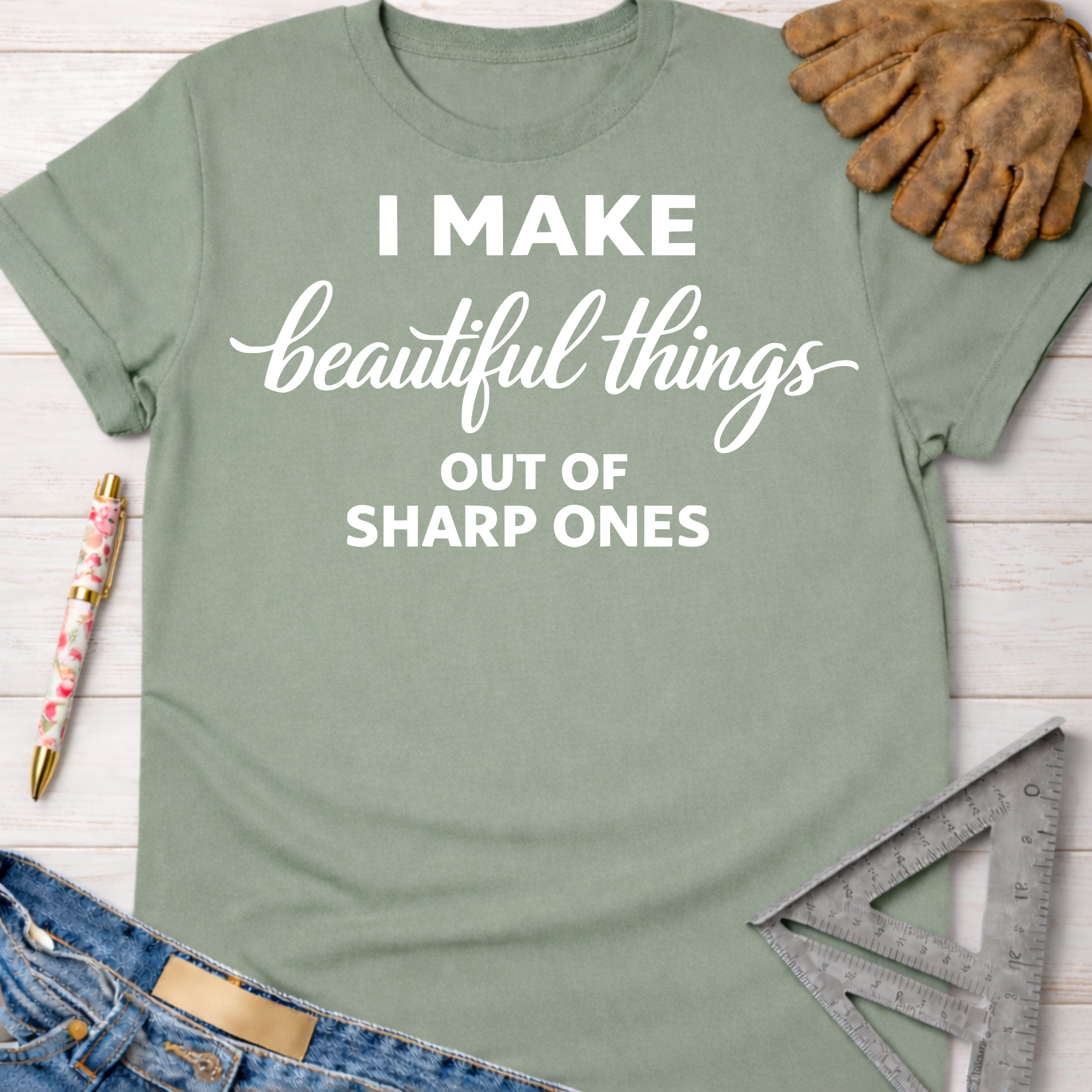Women's funny woodworking graphic tee shirt for female woodworkers, makers, and DIY builders — Sawdust & Ponytails. Sage green cotton tee with "I Make beautiful things OUT OF SHARP ONES" in white text.