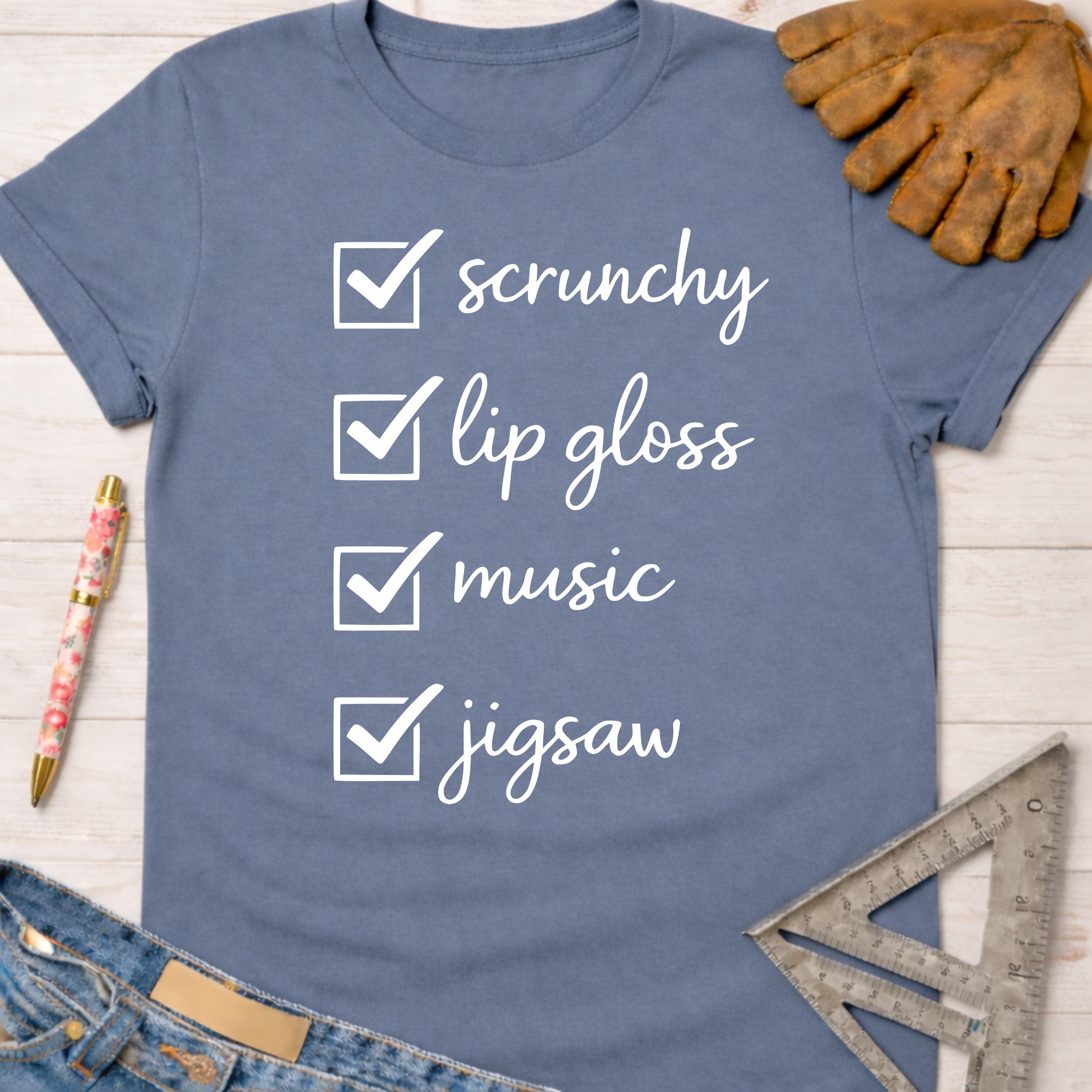 Women's funny woodworking graphic tee shirt for female woodworkers, makers, and DIY builders — Sawdust & Ponytails. Blue crewneck t-shirt with a white "scrunchy, lip gloss, music, jigsaw" checklist design.