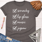 Women's funny woodworking graphic tee shirt for female woodworkers, makers, and DIY builders — Sawdust & Ponytails. Dark gray t-shirt with "scrunchy, lip gloss, music, jigsaw" checklist.