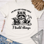 Women's funny woodworking graphic tee shirt for female woodworkers, makers, and DIY builders — Sawdust & Ponytails. White cotton tee with a raccoon holding a drill and hammer, "I'm not like other girls, I build things" text.
