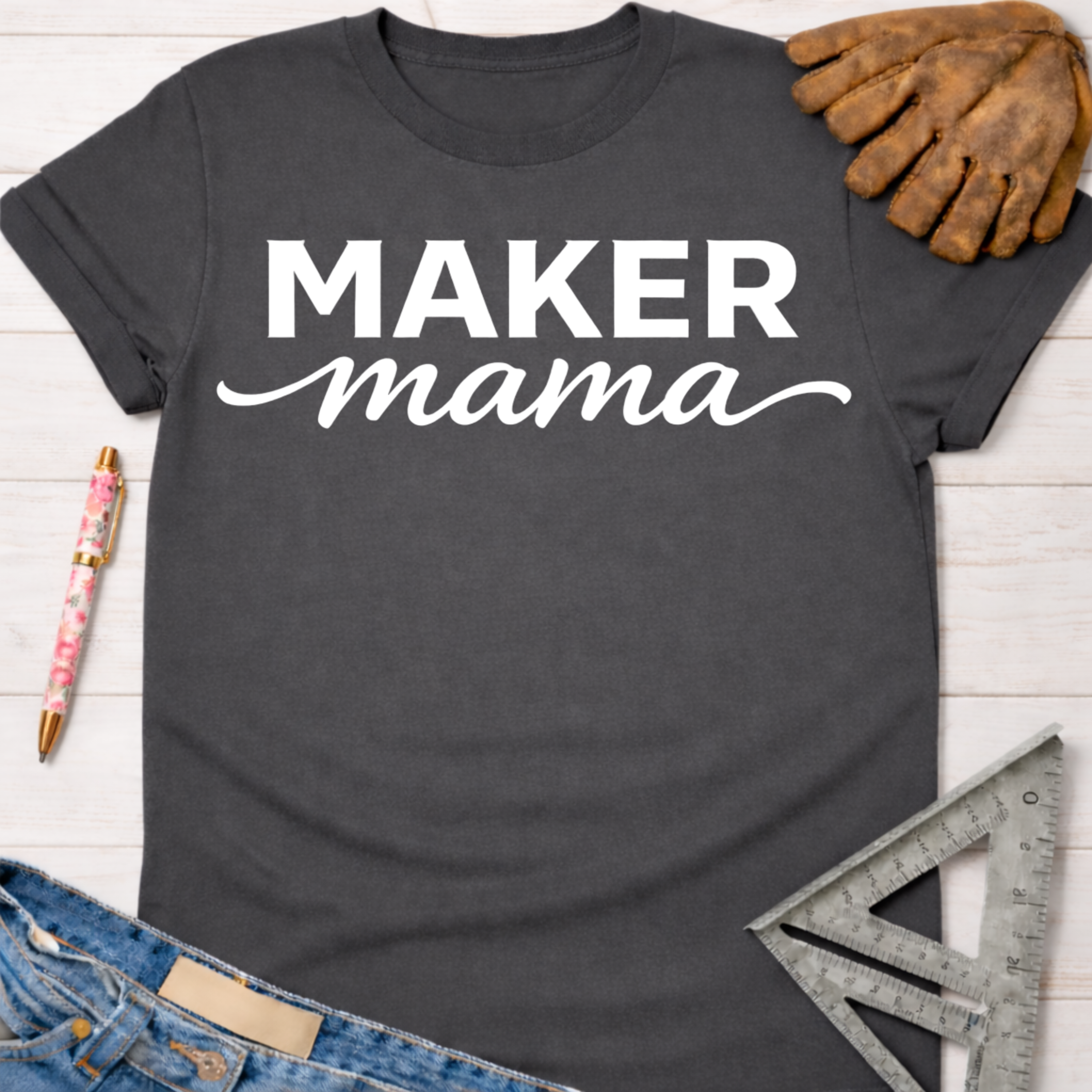 Charcoal gray Maker Mama graphic tee with white text, denim, a pen, and a work glove. Women's funny woodworking graphic tee shirt for female woodworkers, makers, and DIY builders — Sawdust & Ponytails.