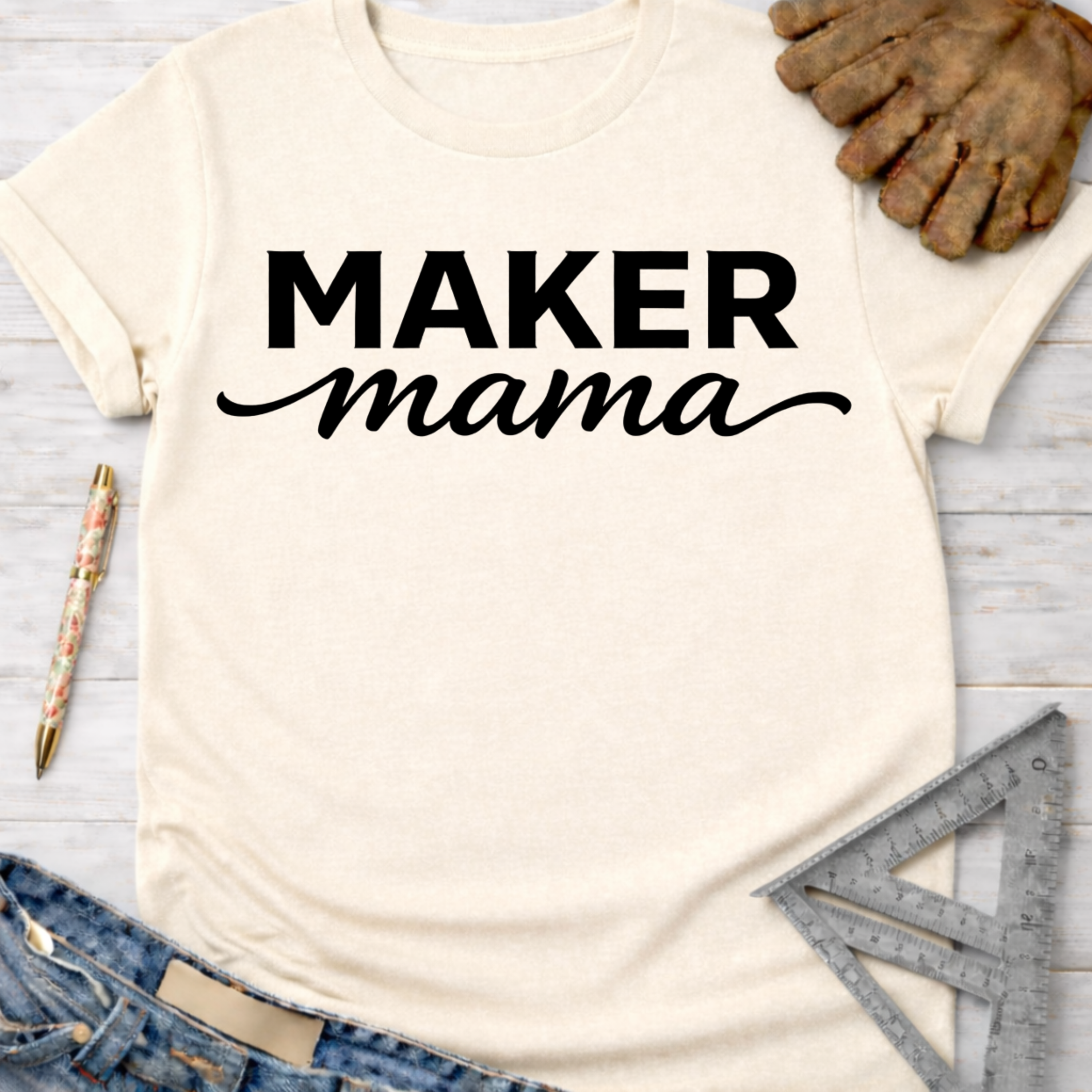 Women's funny woodworking graphic tee shirt for female woodworkers, makers, and DIY builders — Sawdust & Ponytails. Cream "Maker Mama" t-shirt with black text, jeans, work glove, pen, and ruler.