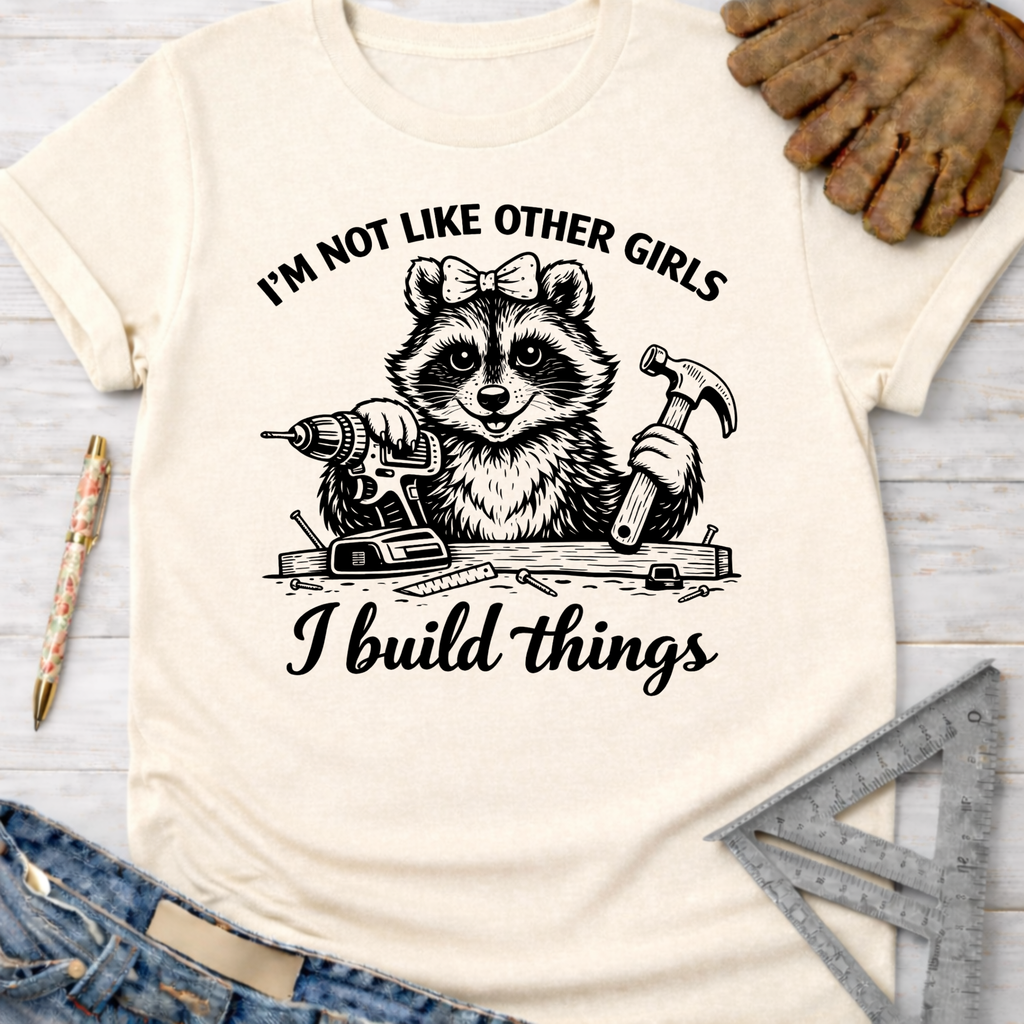 Women's funny woodworking graphic tee shirt for female woodworkers, makers, and DIY builders — Sawdust & Ponytails. Cream t-shirt with a raccoon holding a drill and hammer, text "I'm not like other girls I build things.