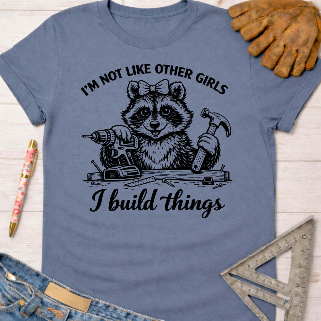 Women's funny woodworking graphic tee shirt for female woodworkers, makers, and DIY builders — Sawdust & Ponytails. Blue cotton tee with a raccoon holding a drill and hammer, text "I'm not like other girls I build things.