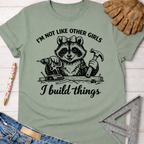 Sage green women's funny woodworking graphic tee with a raccoon holding a drill and hammer, "I'm Not Like Other Girls I Build Things" text. Women's funny woodworking graphic tee shirt for female woodworkers, makers, and DIY builders — Sawdust & Ponytails
