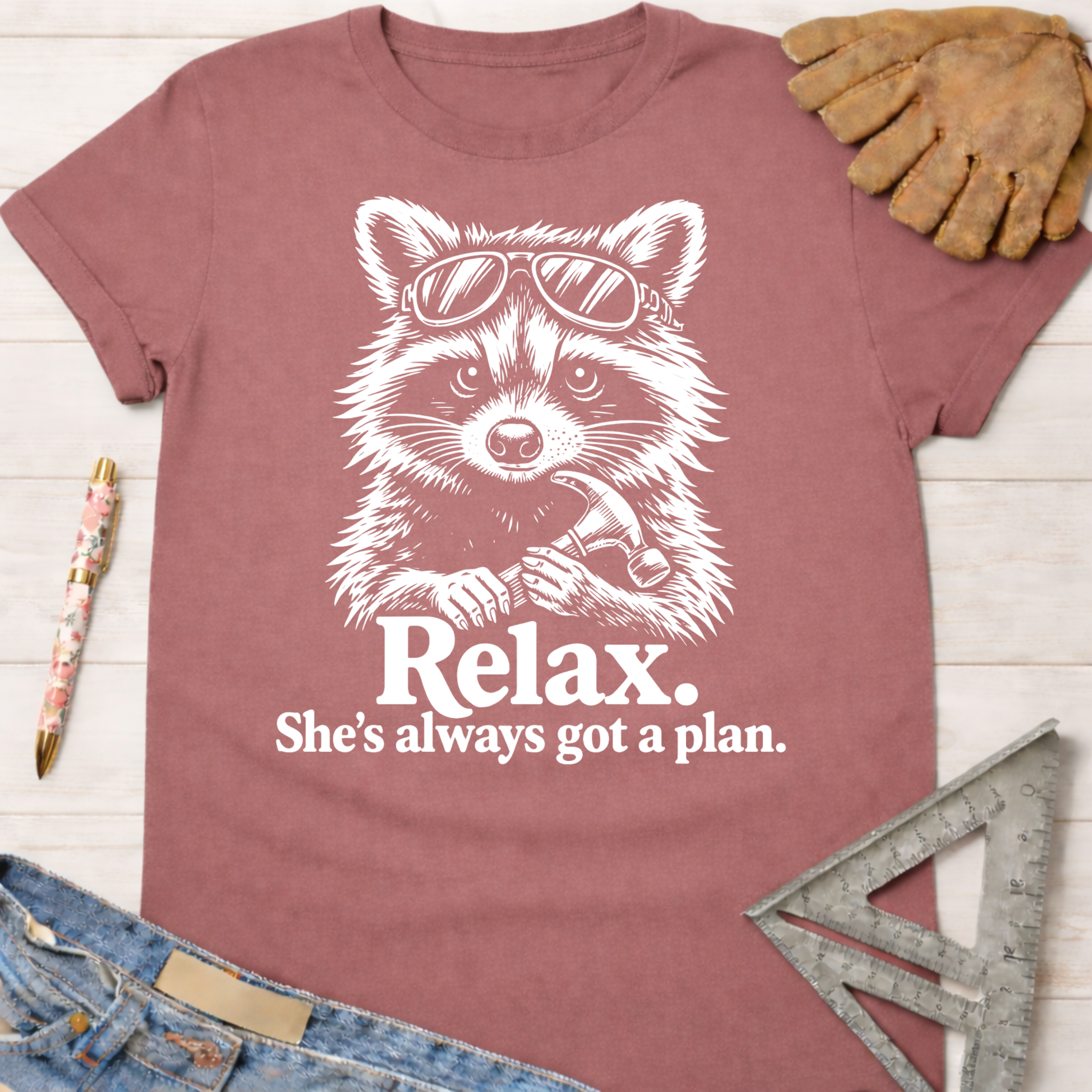 Women's funny woodworking graphic tee shirt for female woodworkers, makers, and DIY builders — Sawdust & Ponytails. Mauve t-shirt with a white graphic of a raccoon wearing sunglasses and holding a hammer, with text "Relax. She's always got a plan.