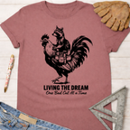 Women's funny woodworking graphic tee shirt for female woodworkers, makers, and DIY builders — Sawdust & Ponytails. Mauve t-shirt with a raccoon riding a rooster, text "Living The Dream One Bad Cut At A Time.