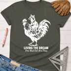 Women's funny woodworking graphic tee shirt for female woodworkers, makers, and DIY builders — Sawdust & Ponytails. Dark gray tee with white graphic of a raccoon riding a rooster, text "LIVING THE DREAM One Bad Cut At a Time.