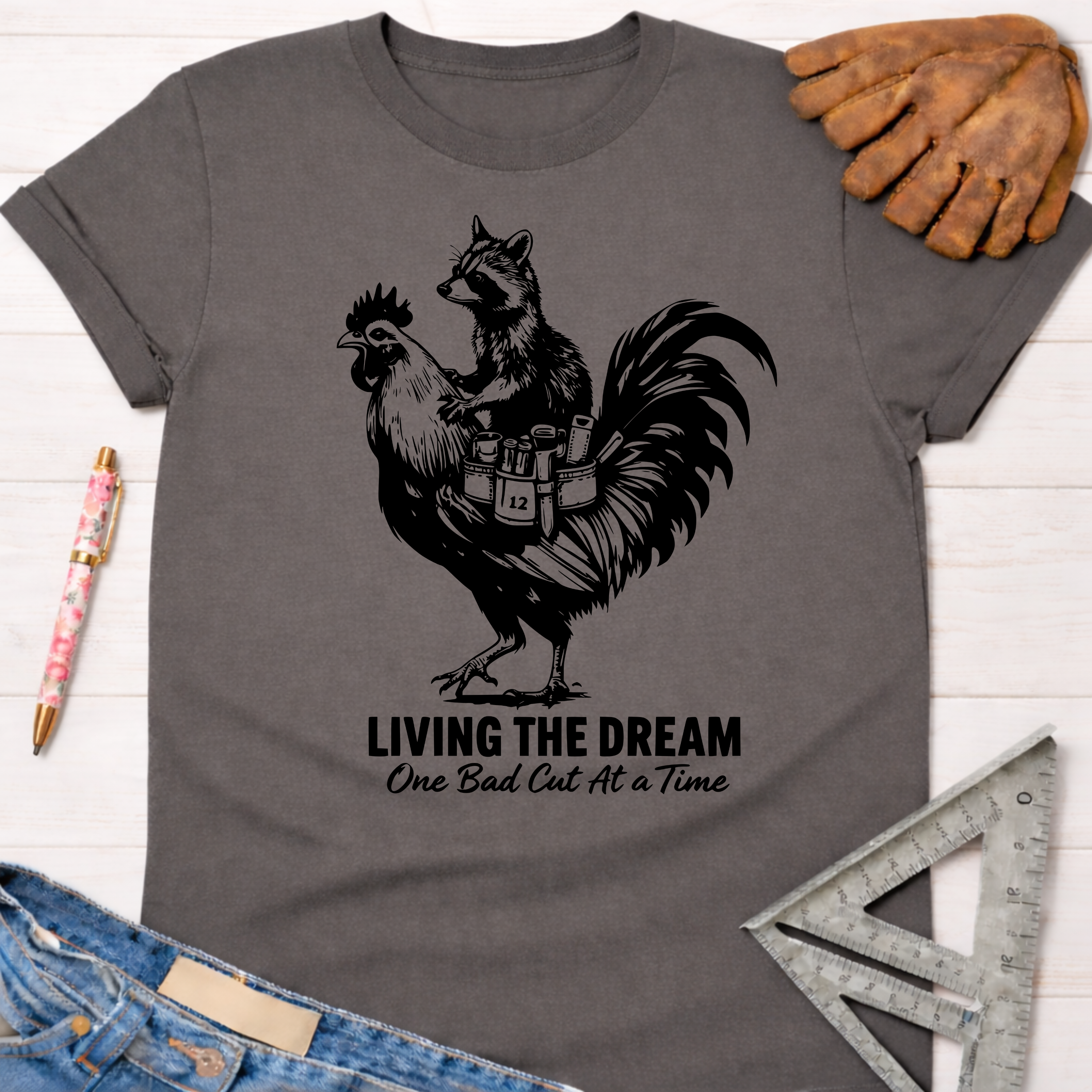 Women's funny woodworking graphic tee shirt for female woodworkers, makers, and DIY builders — Sawdust & Ponytails. Dark gray t-shirt with a black graphic of a raccoon riding a rooster, text "LIVING THE DREAM One Bad Cut At A Time".