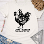 Women's funny woodworking graphic tee shirt for female woodworkers, makers, and DIY builders — Sawdust & Ponytails. White unisex t-shirt with a black graphic of a raccoon riding a rooster, text "LIVING THE DREAM One Bad Cut At a Time".