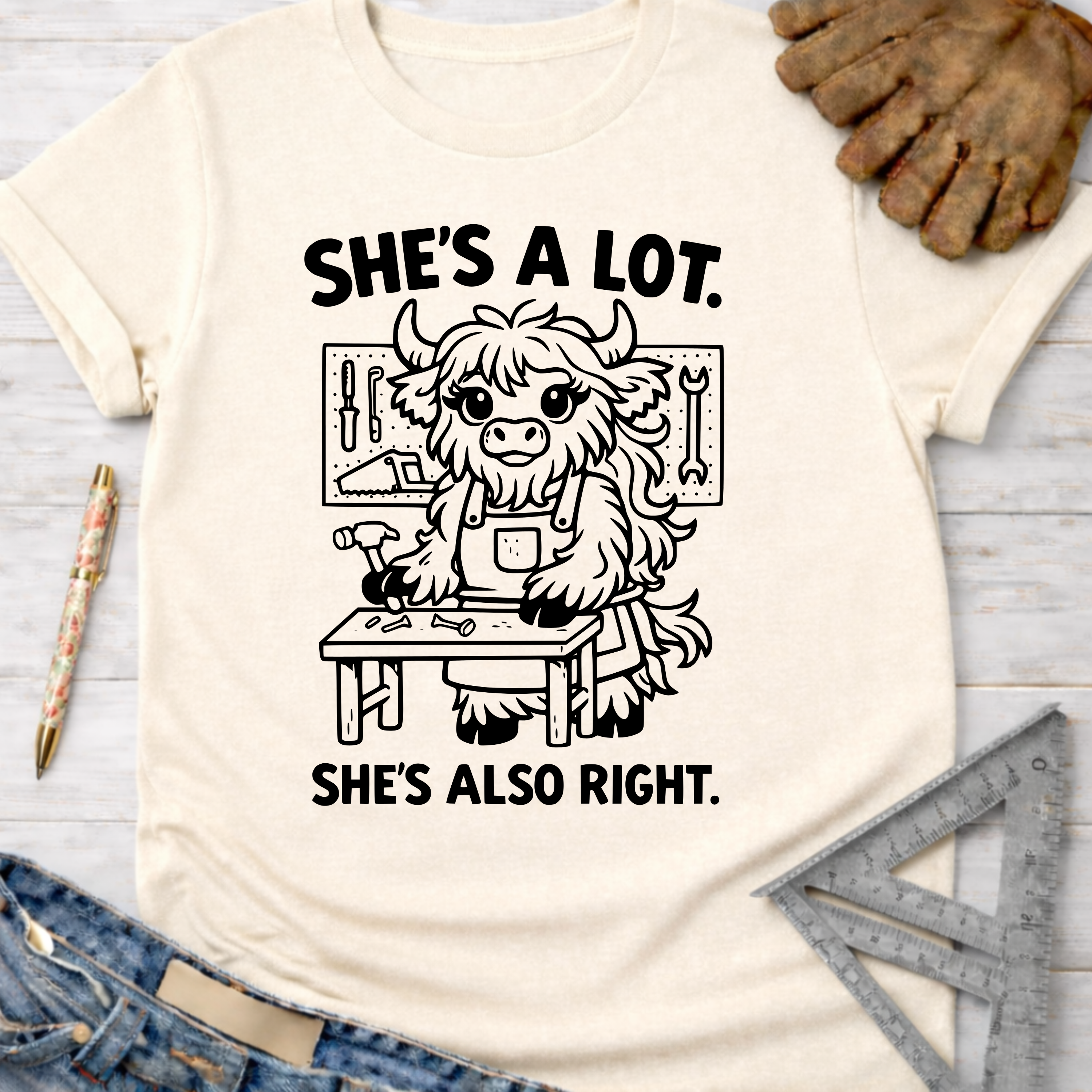 Women's funny woodworking graphic tee shirt for female woodworkers, makers, and DIY builders — Sawdust & Ponytails. Cream t-shirt with "She's A Lot. She's Also Right." text and a cute highland cow working at a workbench.