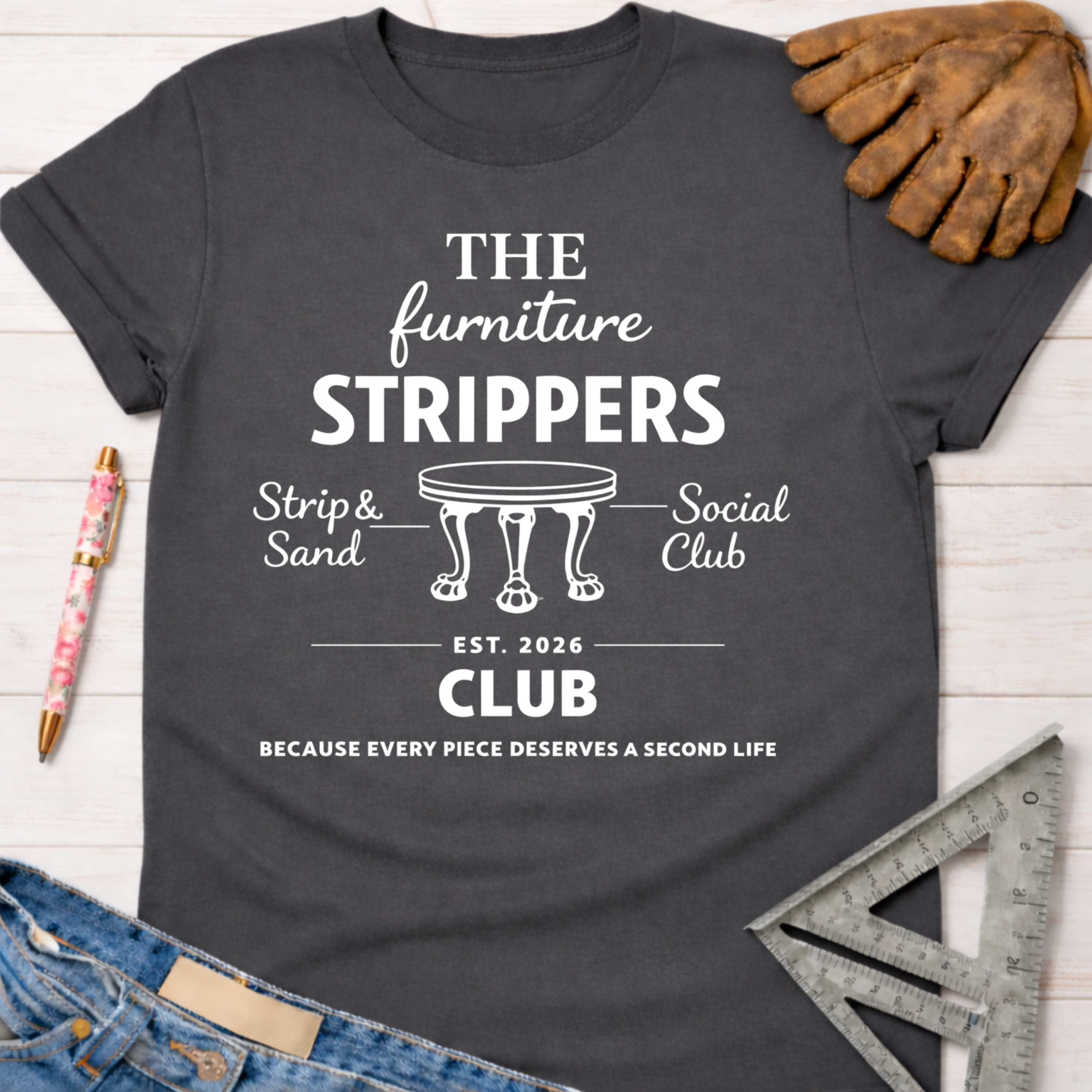 Charcoal gray women's funny woodworking graphic tee with "The Furniture Strippers Social Club" design, jeans, and work glove.