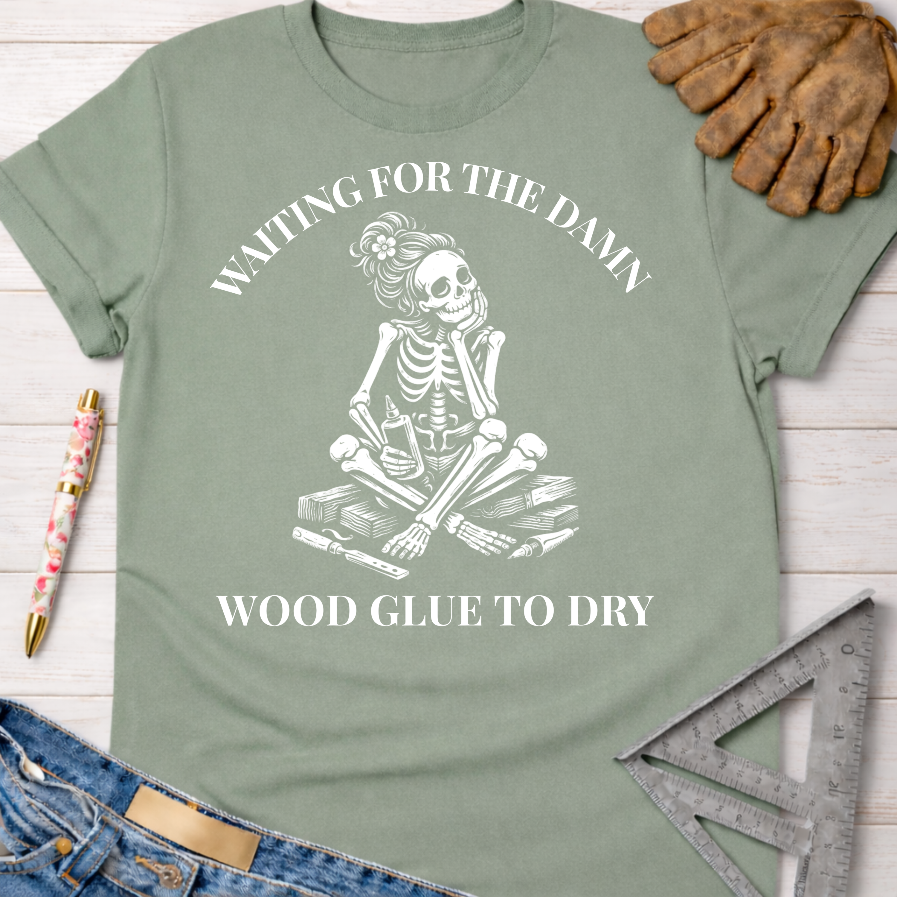 Women's funny woodworking graphic tee shirt for female woodworkers, makers, and DIY builders — Sawdust & Ponytails. Sage green t-shirt with a white skeleton graphic waiting for wood glue to dry.